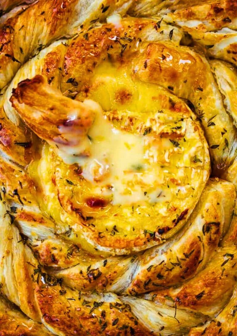 A round puff pastry appetizer with a creamy, melted cheese center, topped with herbs. A piece of bread is dipped into the gooey cheese, surrounded by a golden, twisted pastry crust.