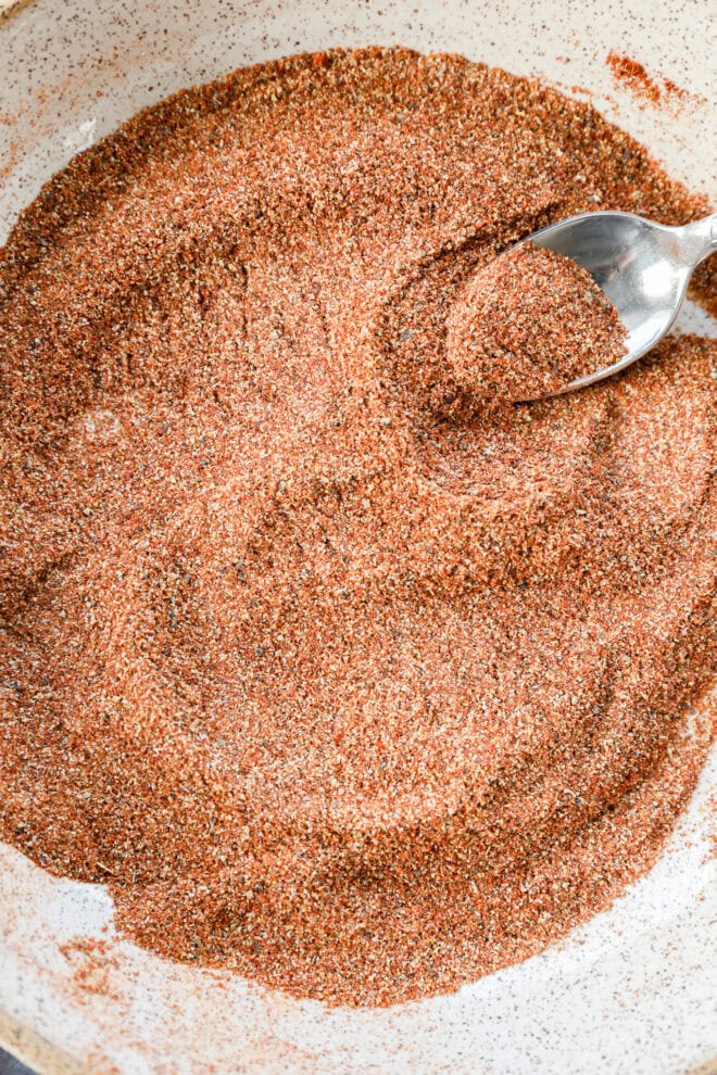 A close-up of a bowl filled with a reddish-brown spice blend, with a metal spoon resting in the mixture and some spices heaped on the spoon.