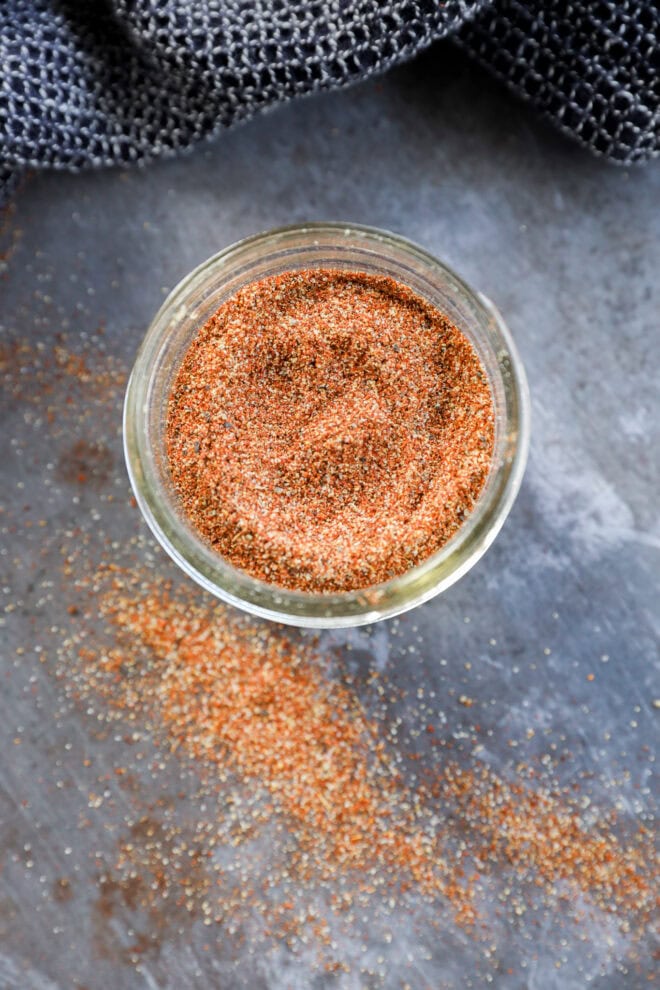 A glass jar filled with a reddish-brown spice blend sits on a gray surface, with some of the fajita seasoning spice scattered around the jar. A textured dark gray cloth is partially visible in the background.