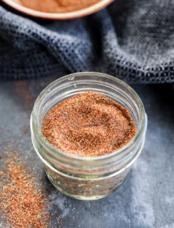 A small glass jar filled with a reddish-brown spice blend sits on a dark surface, with some of the spice spilled nearby and a textured dark cloth and a bowl in the background.