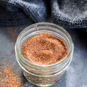 A small glass jar filled with a reddish-brown spice blend sits on a dark surface, with some of the spice spilled nearby and a textured dark cloth and a bowl in the background.