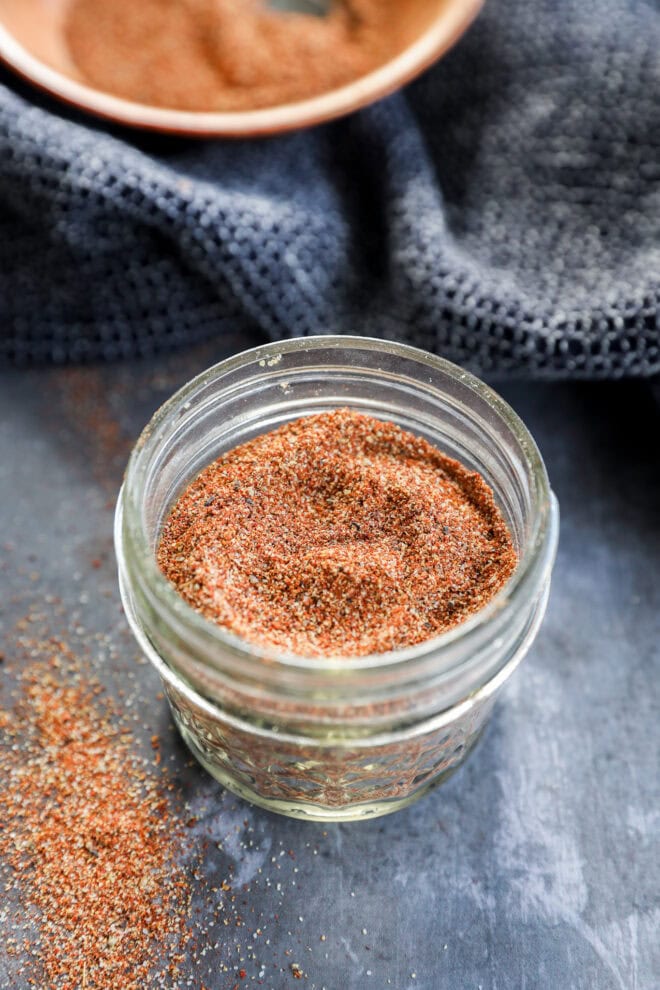 A small glass jar filled with a reddish-brown spice blend sits on a dark surface, with some of the spice spilled nearby and a textured dark cloth and a bowl in the background.