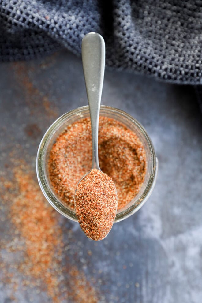 A spoon rests on top of a jar filled with a reddish-brown fajita seasoning blend, with some of the spice spilled on the dark surface below. A textured dark cloth is partially visible in the background.