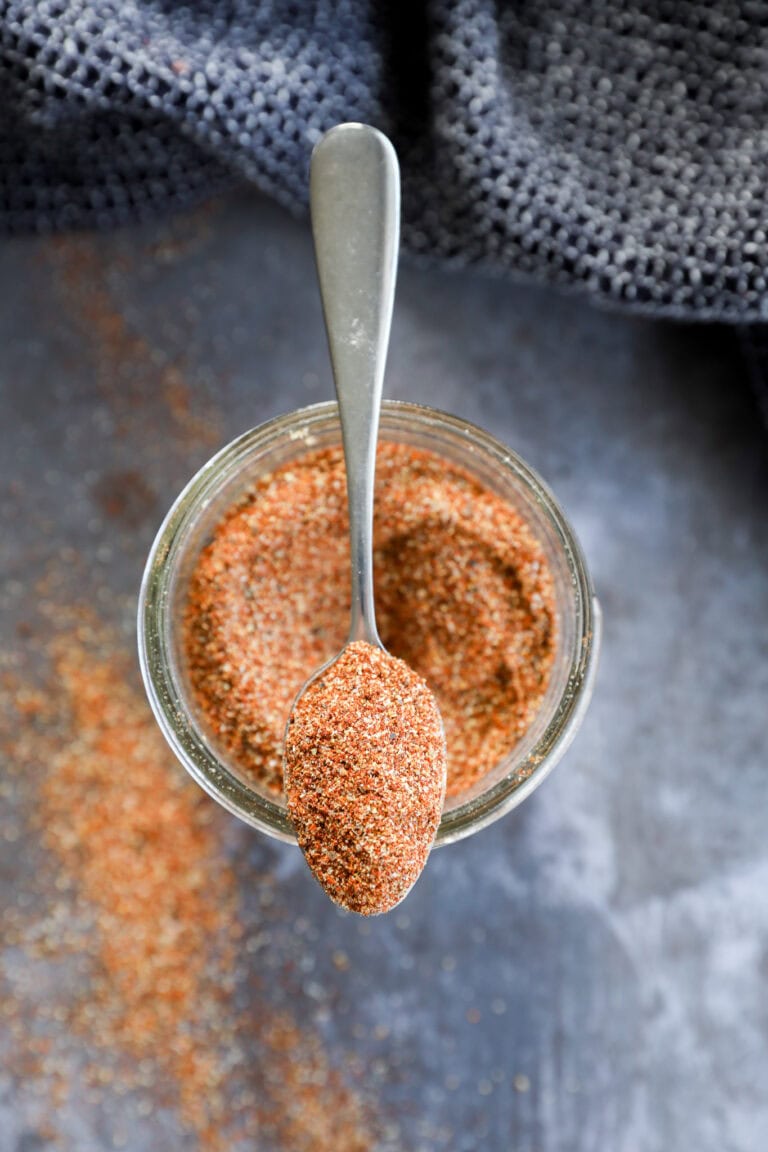 A spoon rests on top of a jar filled with a reddish-brown fajita seasoning blend, with some of the spice spilled on the dark surface below. A textured dark cloth is partially visible in the background.