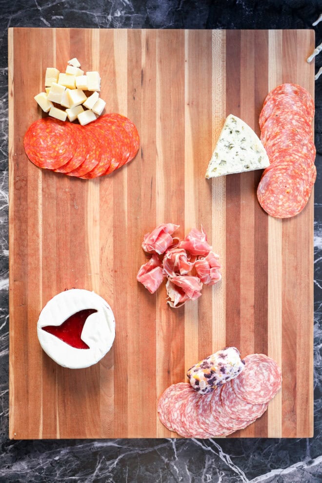 A wooden Halloween charcuterie board features sliced cured meats, cubed cheese, a wedge of blue cheese, a round of brie with jam, a log of goat cheese, and prosciutto artfully arranged on a dark marble surface.