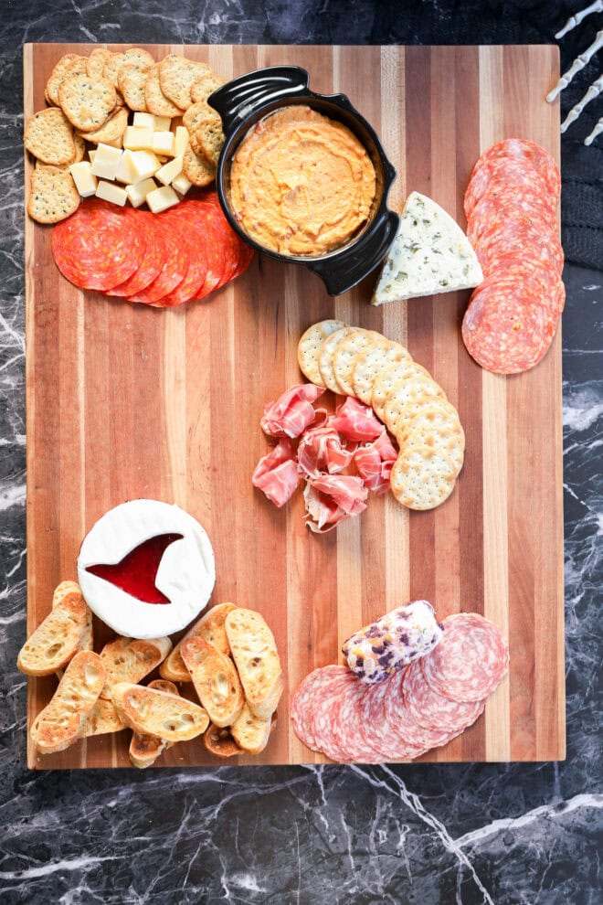 Featuring assorted crackers, cheese cubes, sliced salami, pepperoni, blue cheese, prosciutto, a brie round with jam, a cheese log, crostini, and a bowl of orange dip—perfect for spooky celebrations.