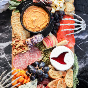 A Halloween charcuterie board featuring meats, cheeses, crackers, fruits, dips, nuts, and bones is artfully arranged with decorative skeleton hands on a dark marble surface for a spooky touch.