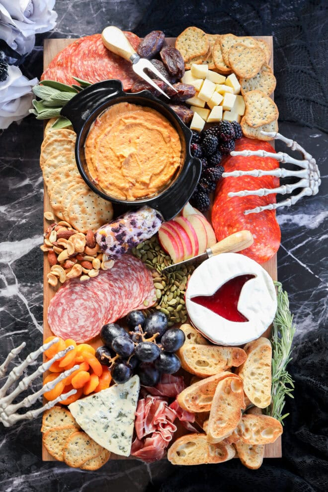 A Halloween charcuterie board featuring meats, cheeses, crackers, fruits, dips, nuts, and bones is artfully arranged with decorative skeleton hands on a dark marble surface for a spooky touch.