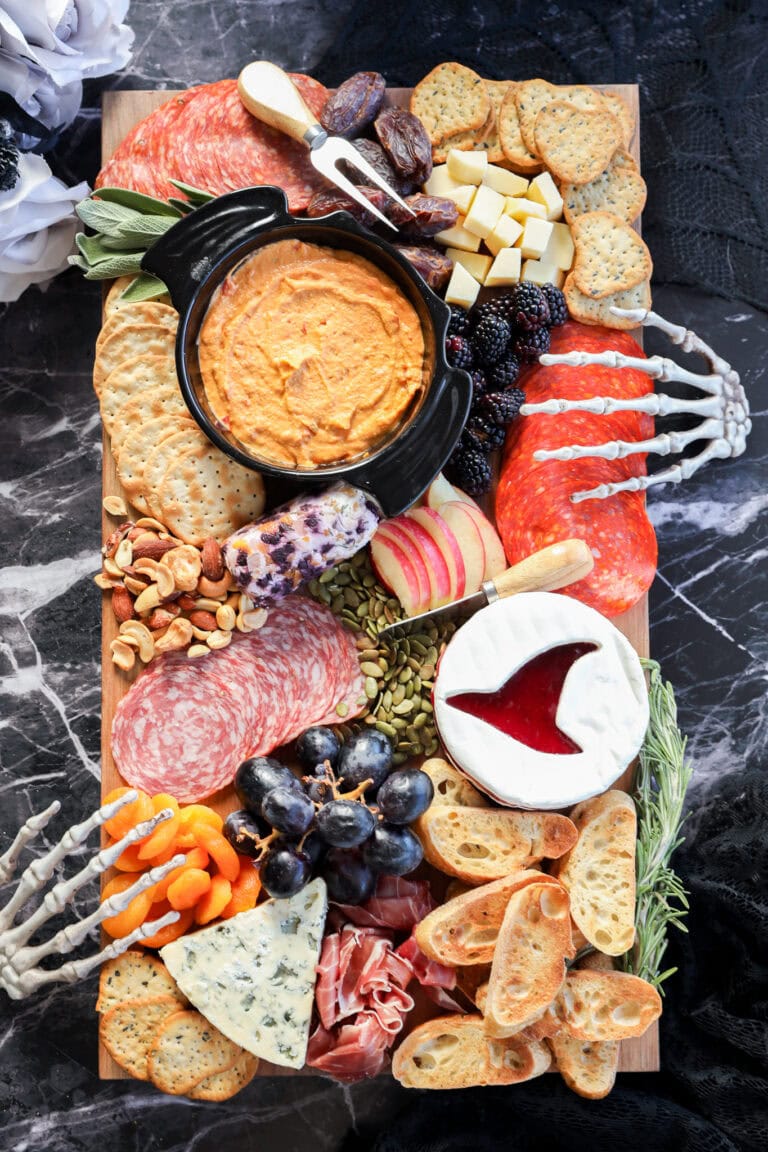 A Halloween charcuterie board featuring meats, cheeses, crackers, fruits, dips, nuts, and bones is artfully arranged with decorative skeleton hands on a dark marble surface for a spooky touch.