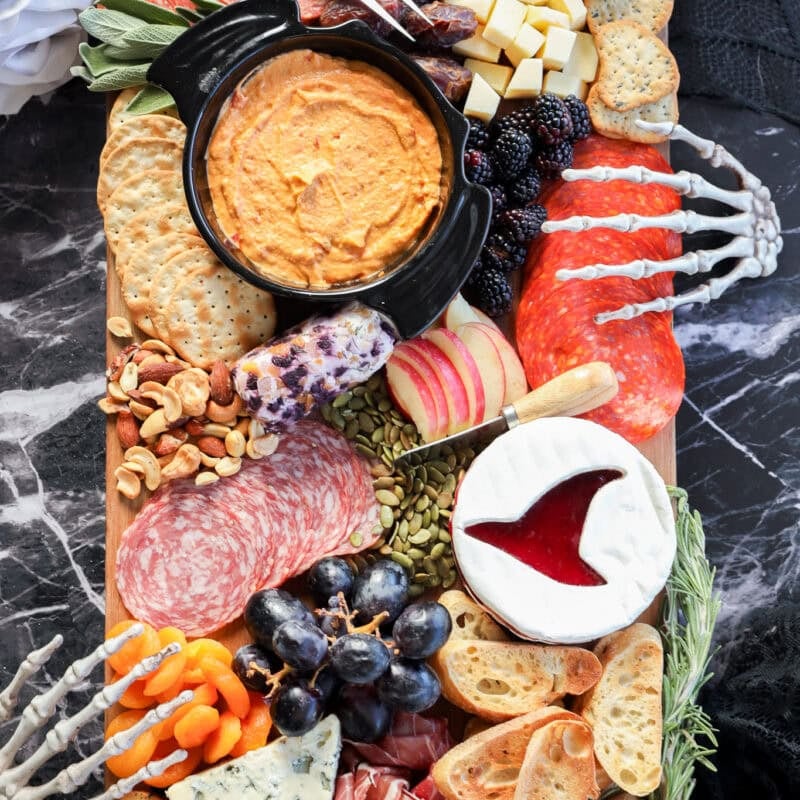 A Halloween charcuterie board featuring meats, cheeses, crackers, fruits, dips, nuts, and bones is artfully arranged with decorative skeleton hands on a dark marble surface for a spooky touch.