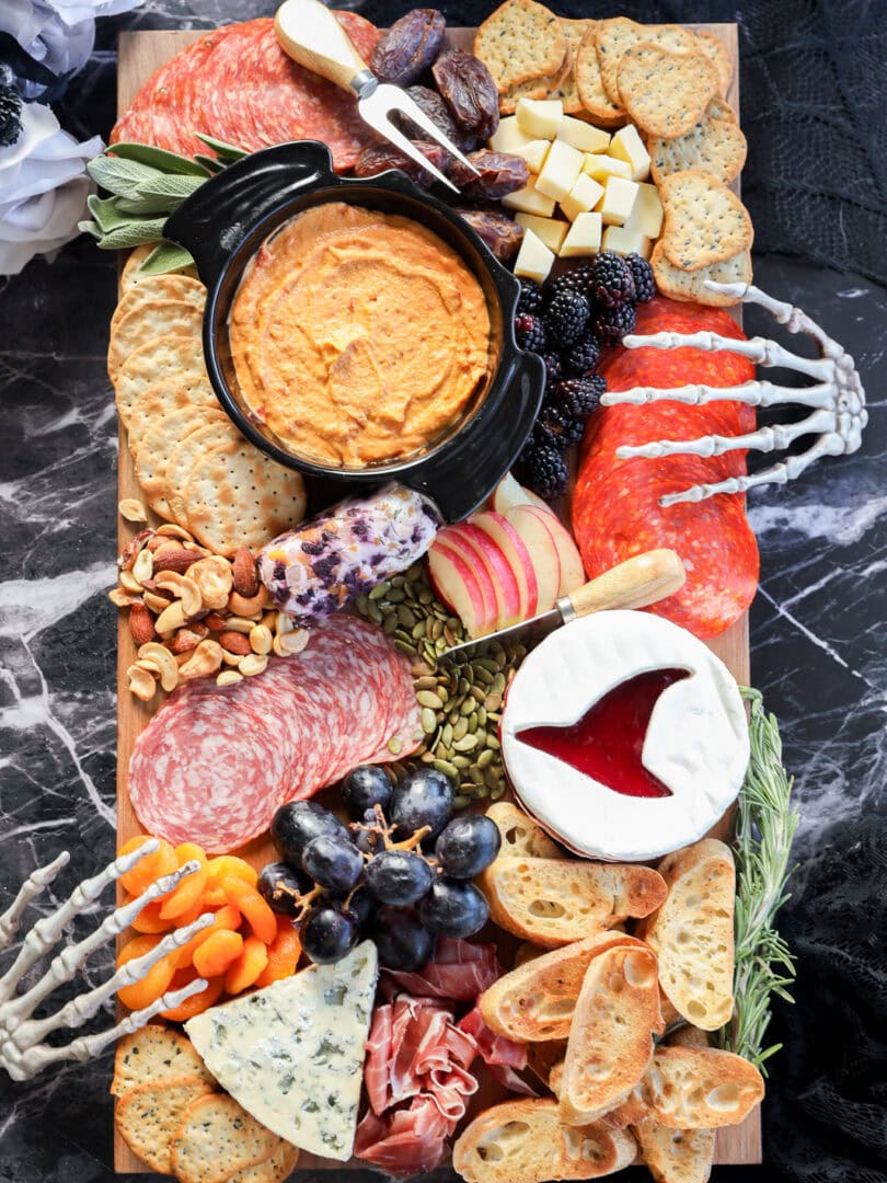 A Halloween charcuterie board featuring meats, cheeses, crackers, fruits, dips, nuts, and bones is artfully arranged with decorative skeleton hands on a dark marble surface for a spooky touch.