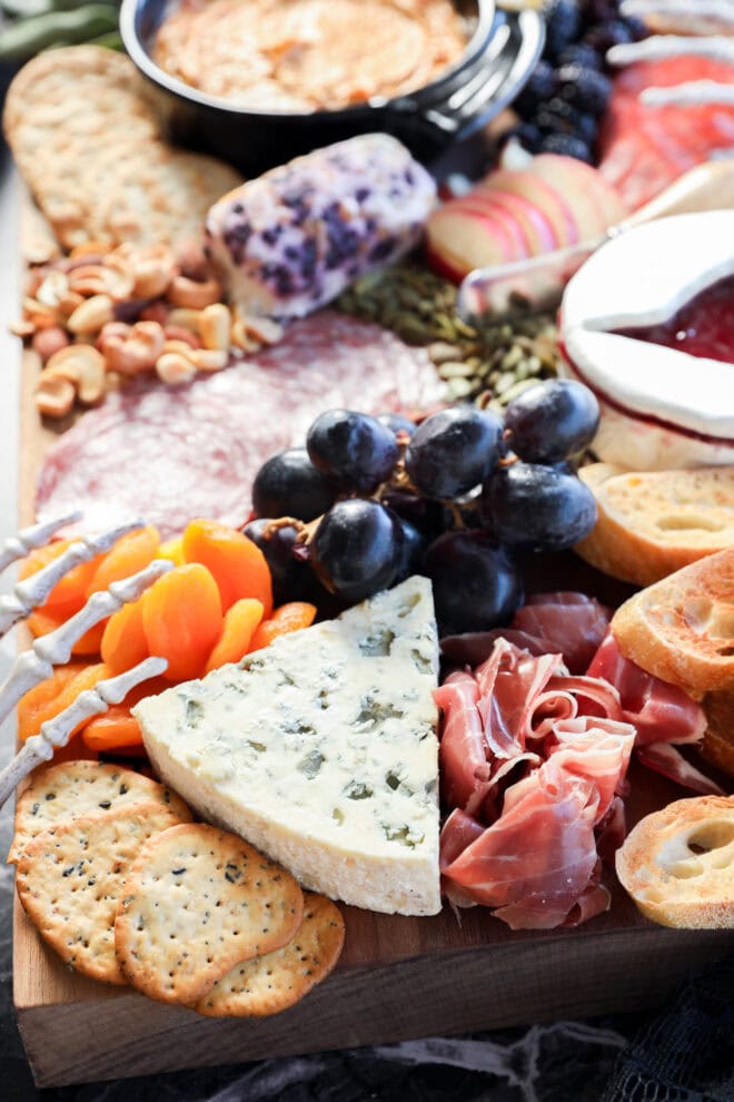 A Halloween charcuterie board featuring crackers, blue cheese, prosciutto, dried apricots, grapes, salami, nuts, sliced bread, a dip in a black bowl, and a small dish of red jam creates an eerie yet delicious spread.