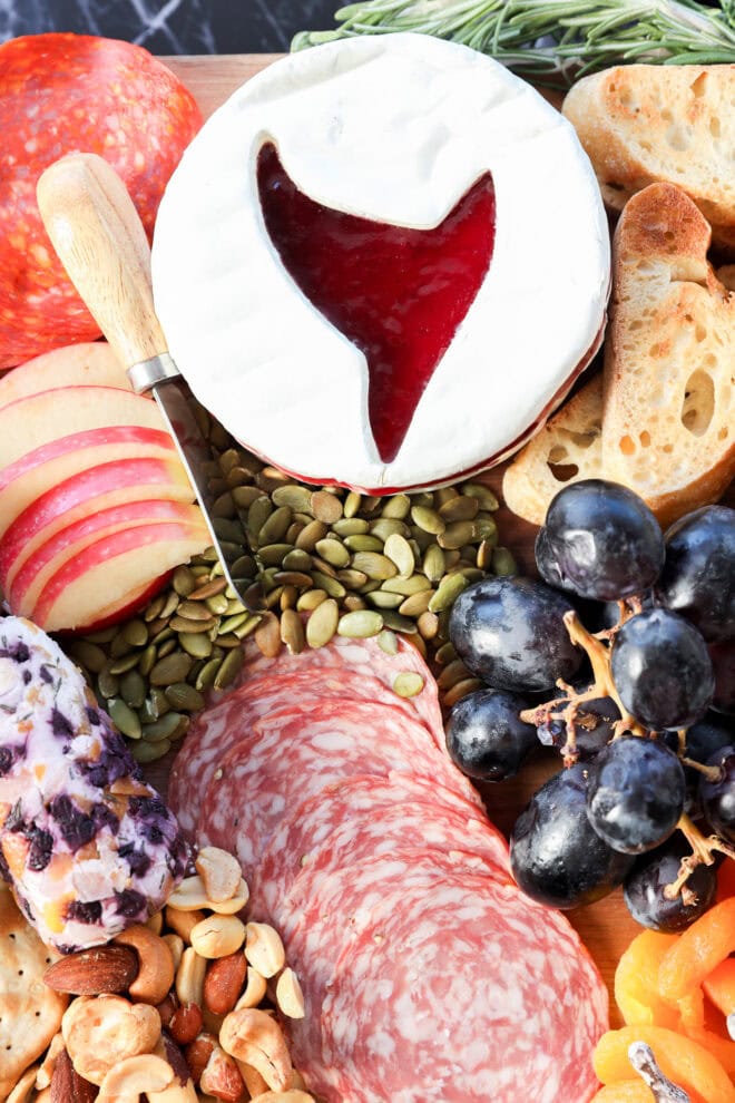 A Halloween charcuterie board featuring sliced meats, apple slices, baguette, grapes, nuts, seeds, crackers, and a round brie cheese topped with red jelly shaped like a whale’s tail. A cheese knife rests nearby.