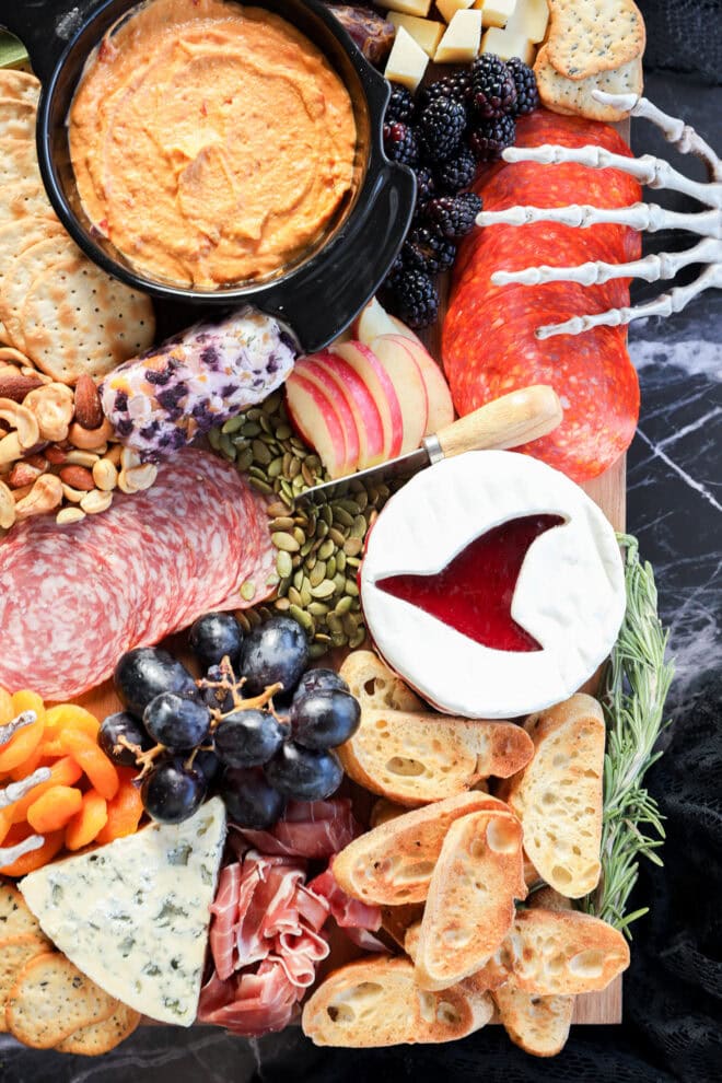 A halloween charcuterie board features cheeses, cured meats, crackers, fruit, nuts, and a brie round topped with red jelly shaped like a whale tail, all arranged beside dip and snacks on a sleek black marble surface.