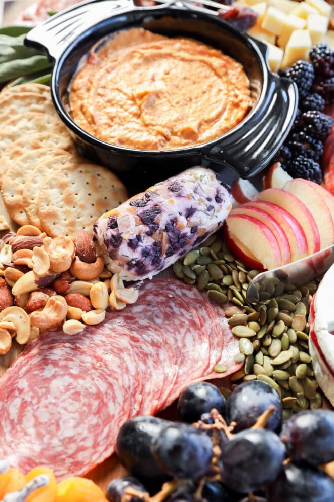 A platter featuring sliced salami, grapes, apple slices, crackers, nuts, pumpkin seeds, cheese, a creamy dip in a black dish, and a log of blueberry goat cheese for a festive touch.