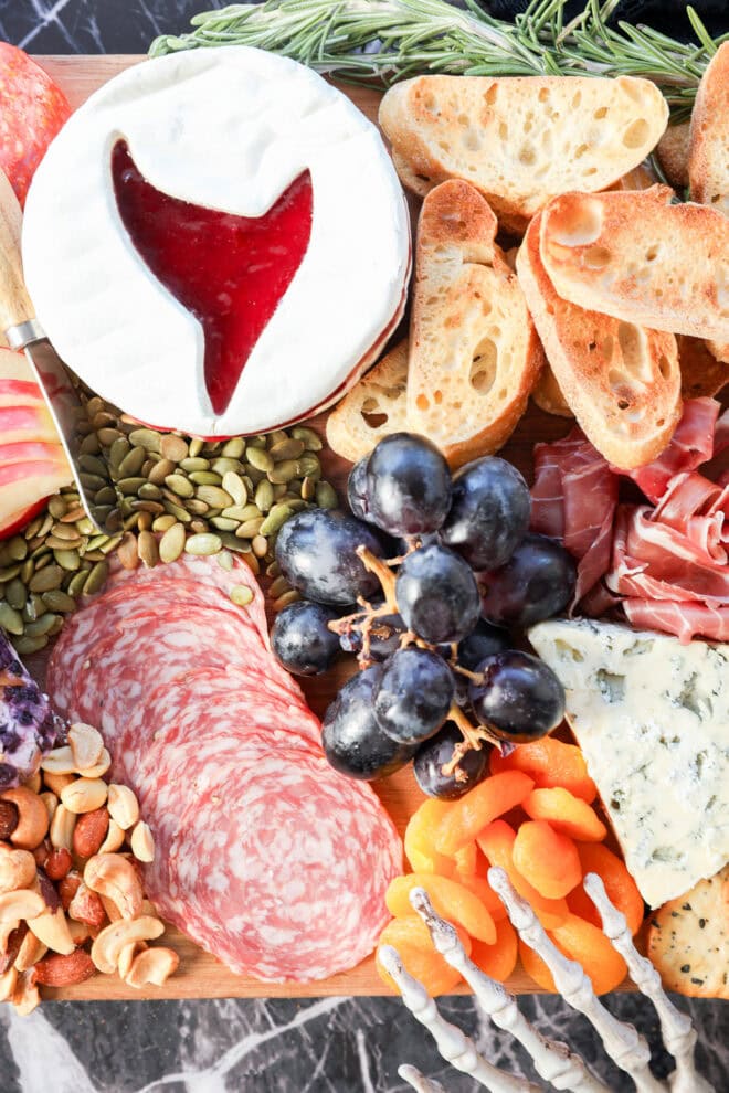 A Halloween charcuterie board featuring sliced bread, brie cheese with a red jam center, salami, prosciutto, blue cheese, black grapes, dried apricots, nuts, seeds, apple slices, and sprigs of rosemary.