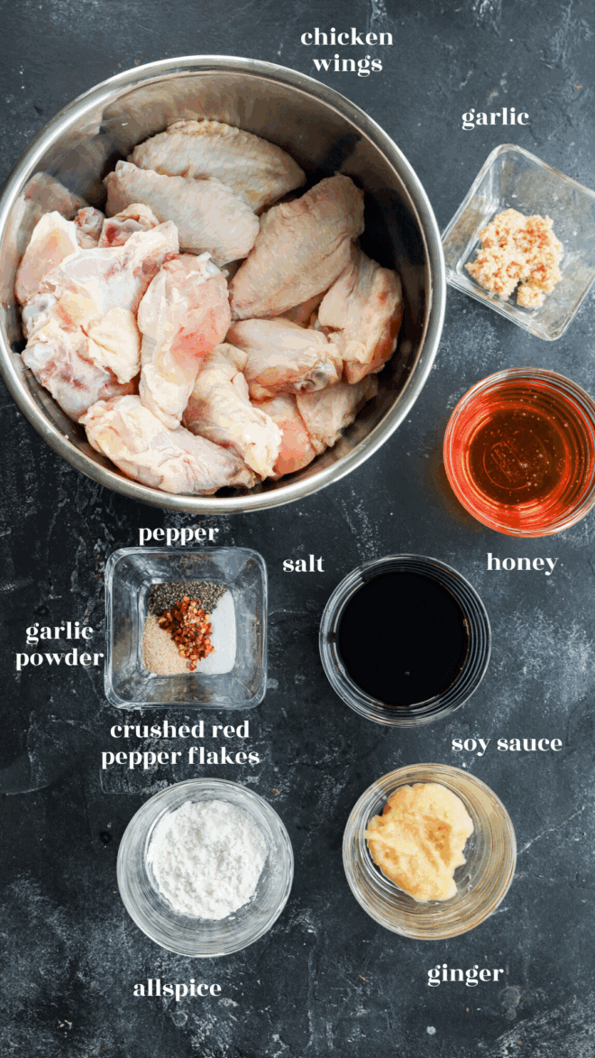 A flat lay of ingredients in bowls on a dark surface: raw chicken wings, minced garlic, honey, soy sauce, ginger, allspice, crushed red pepper flakes, garlic powder, pepper, and salt, each labeled with text. For Honey Garlic Chicken Wings