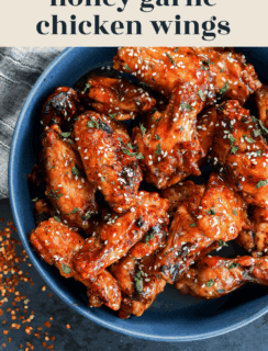 A blue bowl filled with glazed honey garlic chicken wings, sprinkled with sesame seeds and chopped herbs, sits on a dark surface. Text above reads honey garlic chicken wings.
