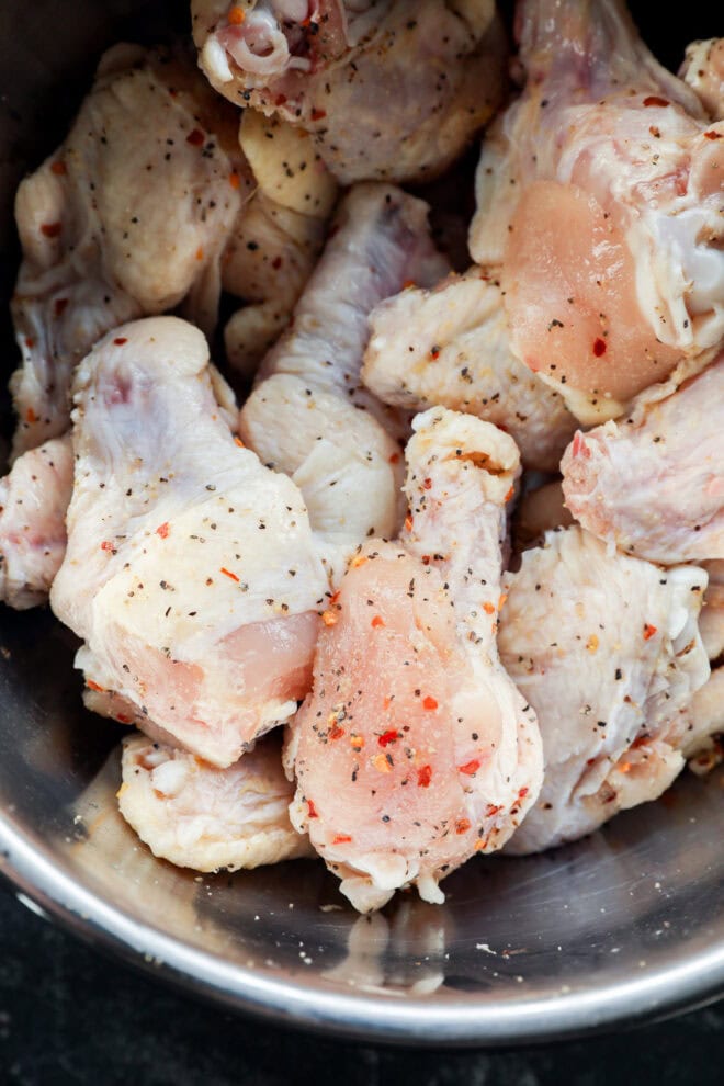 A close-up of raw chicken drumsticks and wings in a metal bowl, seasoned with black pepper and red chili flakes.