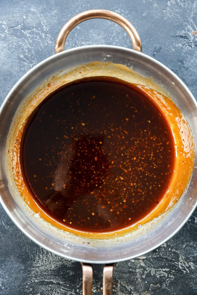 A saucepan filled with a dark brown sauce, likely caramel or soy-based, sits on a textured gray surface. The sauce has a glossy appearance with small specks throughout and some residue on the pans sides.