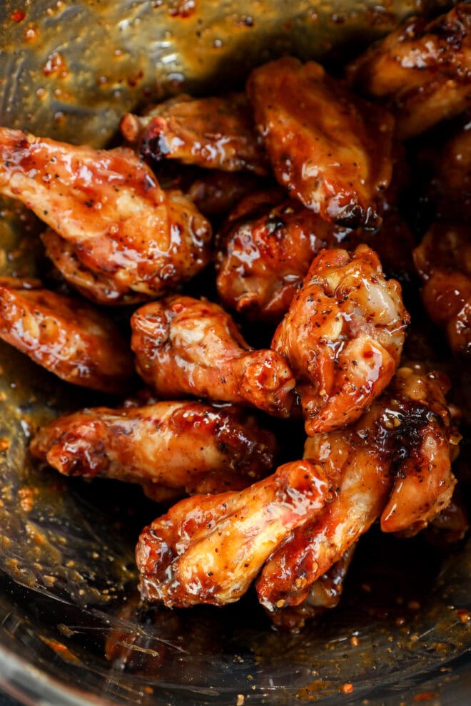 A close-up of glazed honey garlic chicken wings coated in a sticky, reddish-brown sauce with visible seasoning, piled together in a glass bowl.