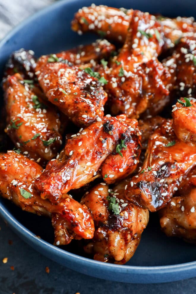 A blue bowl filled with glazed chicken wings, garnished with sesame seeds and chopped herbs. The wings appear caramelized and saucy, giving them a glossy and appetizing look.