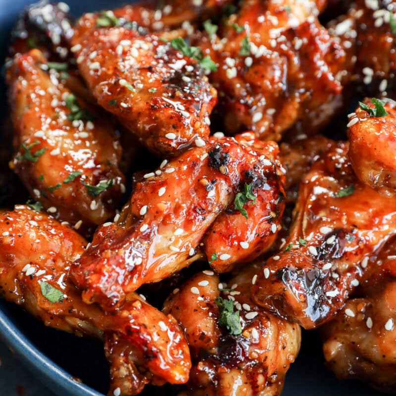 A blue bowl filled with glazed chicken wings, garnished with sesame seeds and chopped herbs. The wings appear caramelized and saucy, giving them a glossy and appetizing look.