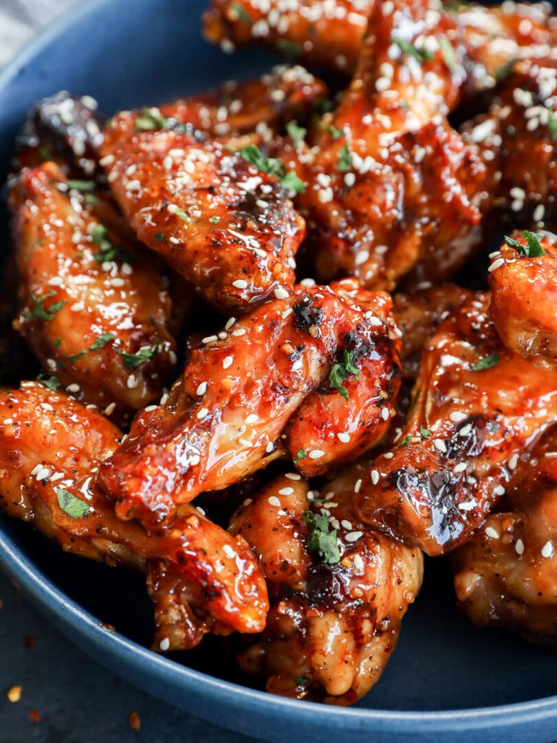 A blue bowl filled with glazed chicken wings, garnished with sesame seeds and chopped herbs. The wings appear caramelized and saucy, giving them a glossy and appetizing look.