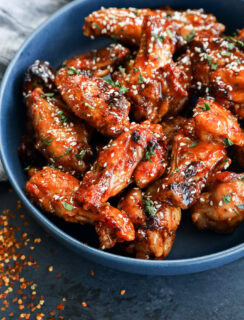 A blue bowl filled with glazed chicken wings sprinkled with sesame seeds and chopped herbs, with some red pepper flakes scattered on a dark surface nearby.