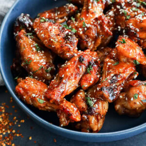 A blue bowl filled with glazed chicken wings sprinkled with sesame seeds and chopped herbs, with some red pepper flakes scattered on a dark surface nearby.