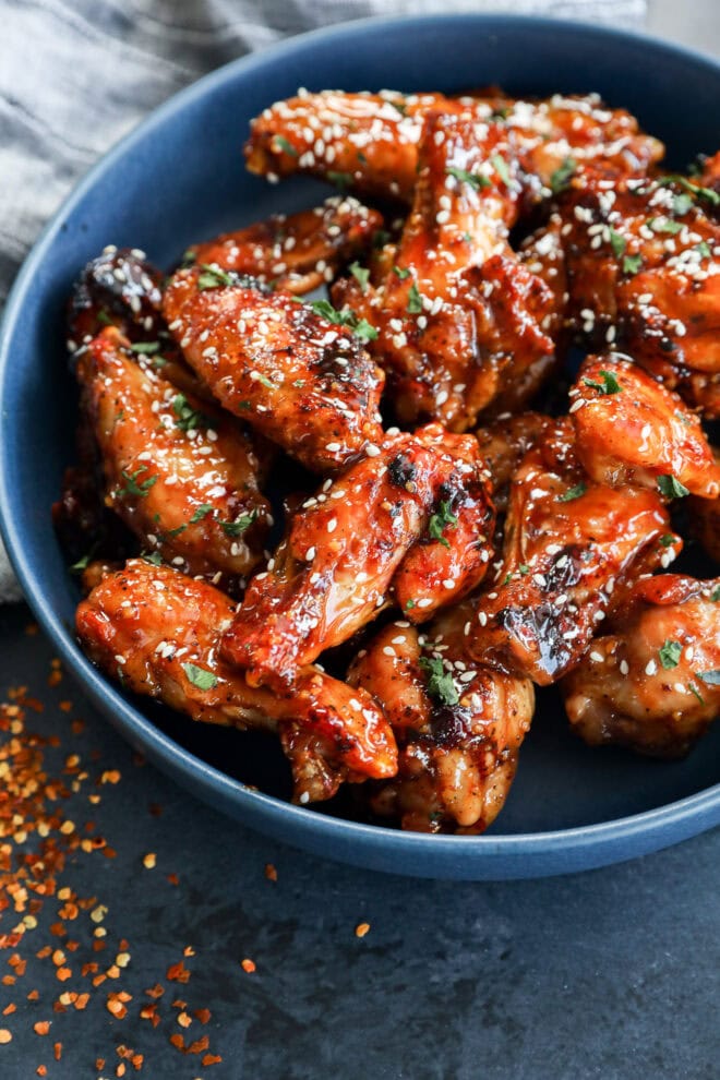 A blue bowl filled with glazed chicken wings sprinkled with sesame seeds and chopped herbs, with some red pepper flakes scattered on a dark surface nearby.