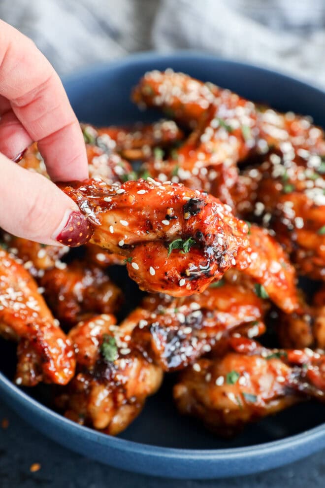A close-up of a hand picking up a glazed chicken wing from a blue bowl filled with sticky, saucy wings garnished with white sesame seeds and chopped herbs.