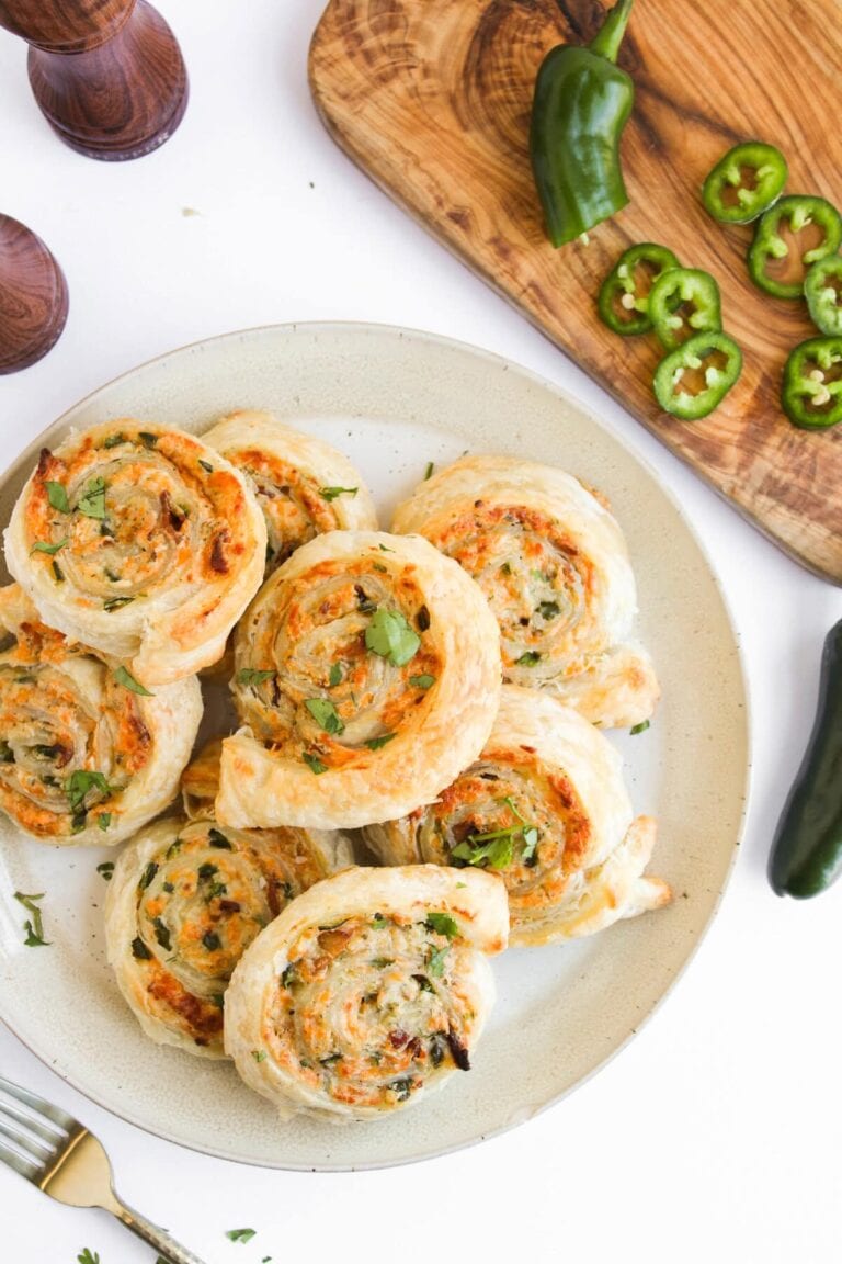 A plate of golden, flaky puff pastry appetizers pinwheels garnished with herbs sits on a white surface. Nearby, sliced jalapeños and whole peppers rest on a wooden cutting board.