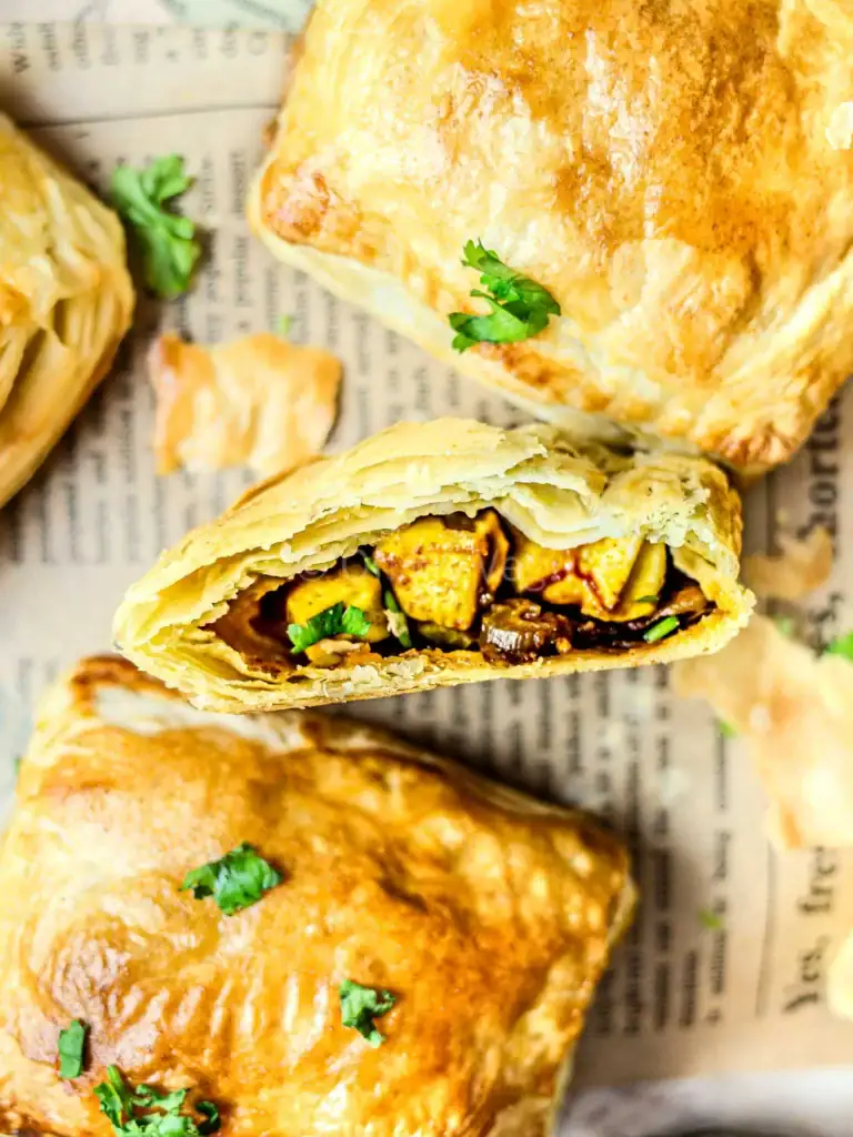 Golden, flaky puff pastry appetizers filled with spicy paneer cubes and vegetables, garnished with fresh cilantro, displayed on a sheet of newspaper for a rustic presentation.