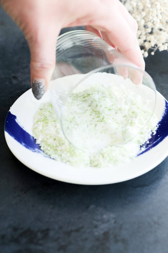 A hand rolls the rim of a glass in salt and lime zest on a white plate with a blue stripe, prepping the glass.