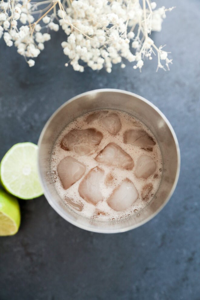 A metal cup filled with ice and a fizzy Prickly Pear Margarita sits on a dark surface, next to two lime halves and white dried flowers.