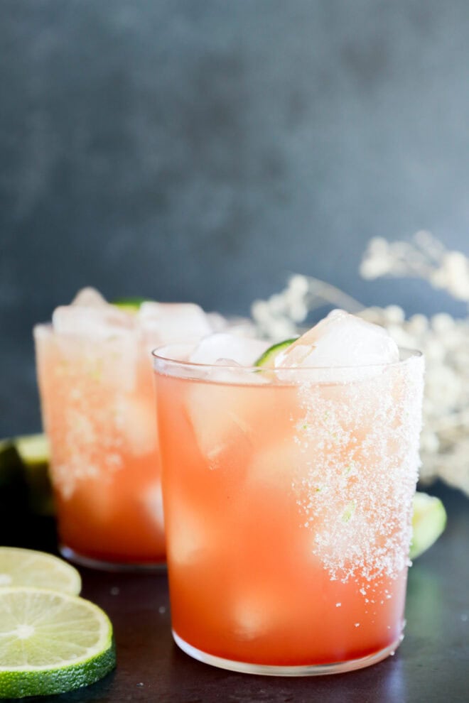 Two glasses with ice, salted rims, and lime wedges sit on a dark surface, surrounded by lime slices and blurred white flowers in the background.