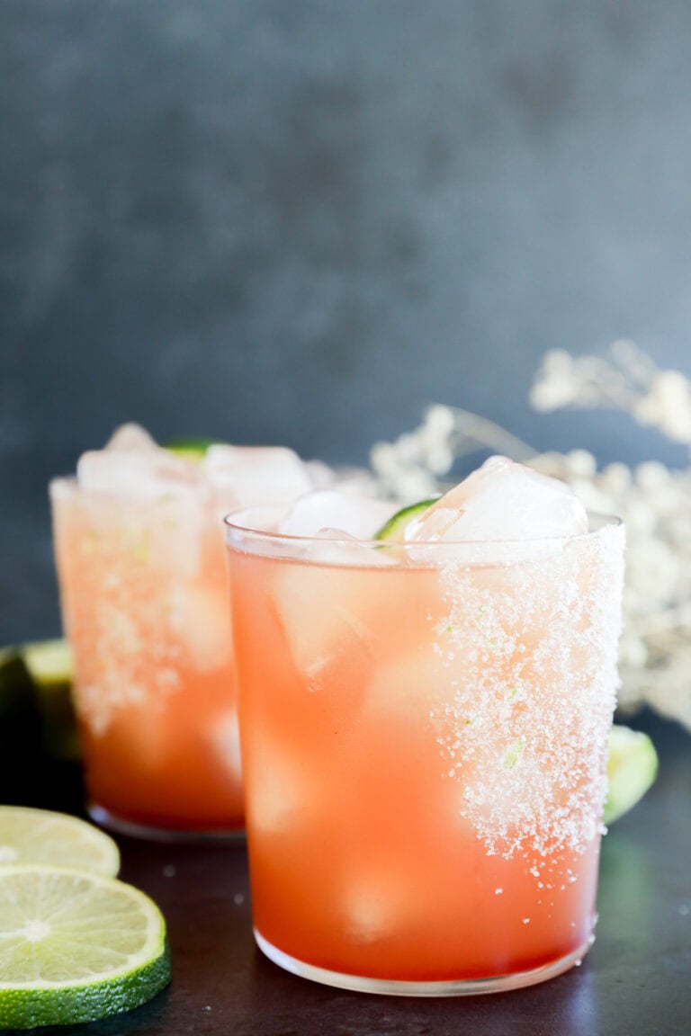 Two glasses with ice, salted rims, and lime wedges sit on a dark surface, surrounded by lime slices and blurred white flowers in the background.