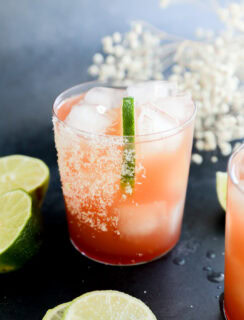 A glass filled with a vibrant Prickly Pear Margarita and ice cubes, garnished with a lime wedge and a salted rim. Slices of lime are scattered on the dark surface around the glass, with white flowers blurred in the background.