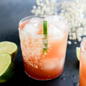 A glass filled with a vibrant Prickly Pear Margarita and ice cubes, garnished with a lime wedge and a salted rim. Slices of lime are scattered on the dark surface around the glass, with white flowers blurred in the background.