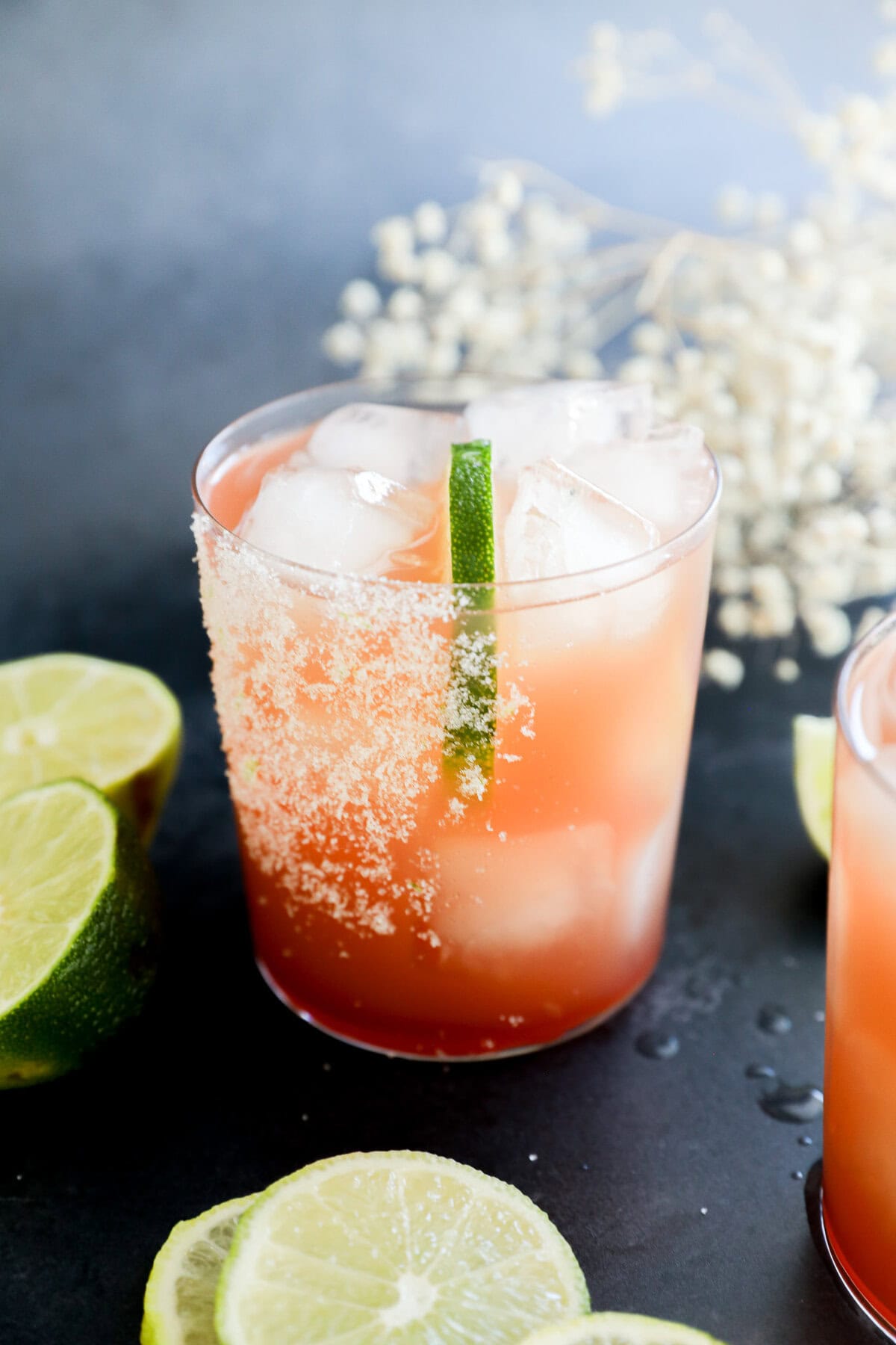 A glass filled with a vibrant Prickly Pear Margarita and ice cubes, garnished with a lime wedge and a salted rim. Slices of lime are scattered on the dark surface around the glass, with white flowers blurred in the background.