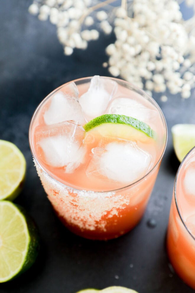 A glass filled with a pinkish-orange drink over ice, garnished with a lime wedge and a salted rim. Slices of lime and white flowers are visible in the background on a dark surface.