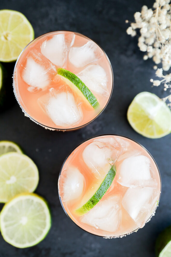 Two glasses filled with a pink Prickly Pear Margarita, ice cubes, and a lime wedge, surrounded by sliced limes on a dark surface.