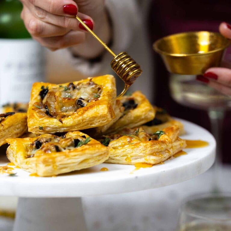 A hand drizzles honey onto puff pastry appetizers topped with cheese and herbs, displayed on a white cake stand. Another hand holds a small golden honey pot nearby.