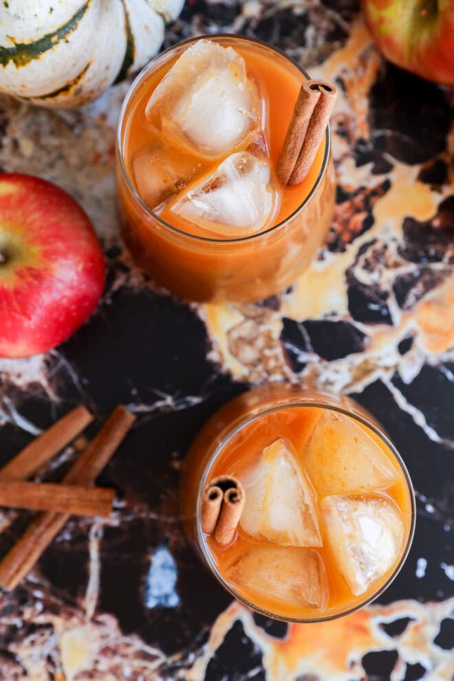 Two glasses of iced pumpkin juice, garnished with cinnamon sticks, sit on a marbled surface surrounded by apples, a small white pumpkin, and loose cinnamon sticks.