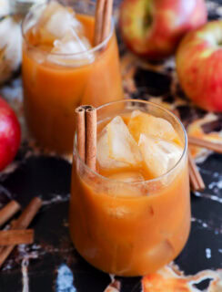 Two glasses filled with pumpkin juice, garnished with cinnamon sticks, sit on a dark marble surface surrounded by red apples and extra cinnamon sticks.