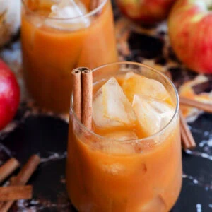 Two glasses filled with pumpkin juice, garnished with cinnamon sticks, sit on a dark marble surface surrounded by red apples and extra cinnamon sticks.