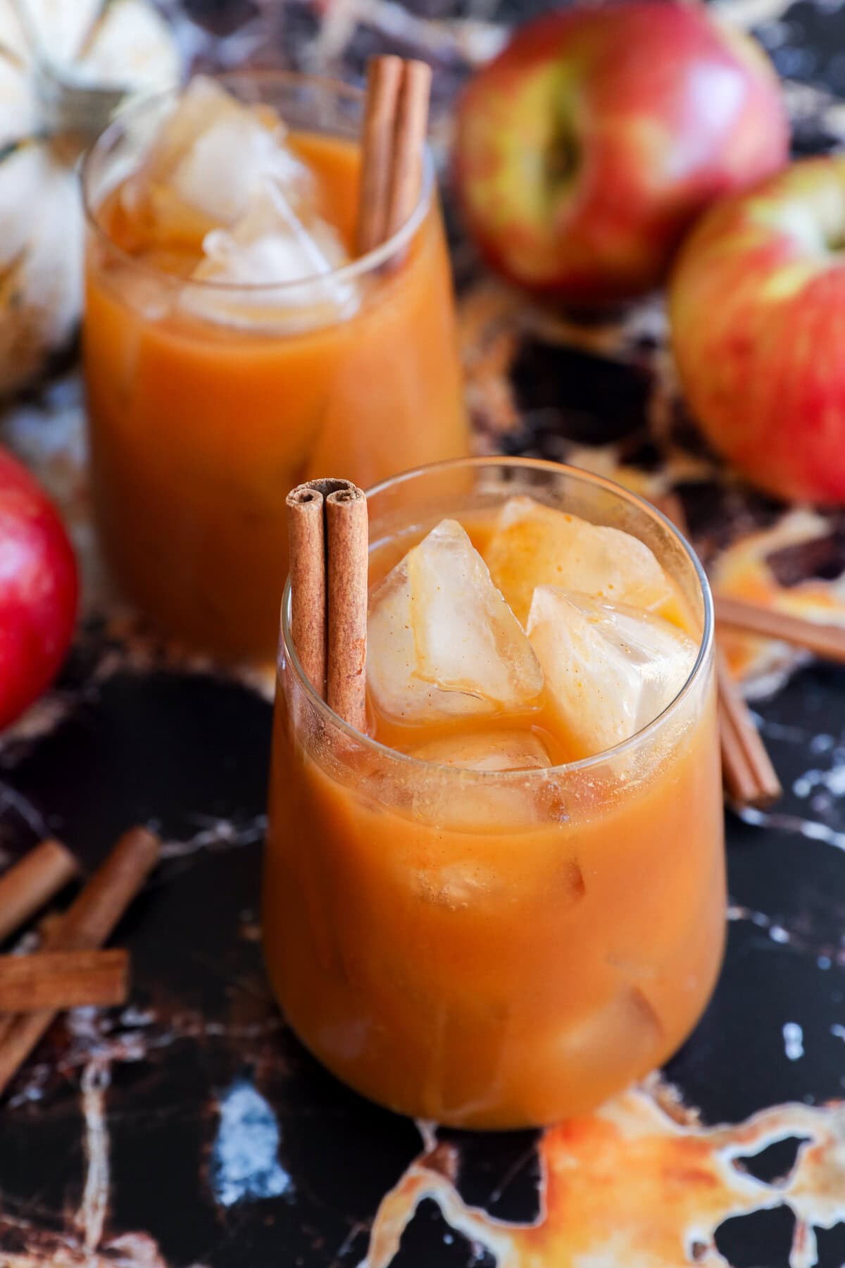 Two glasses filled with pumpkin juice, garnished with cinnamon sticks, sit on a dark marble surface surrounded by red apples and extra cinnamon sticks.