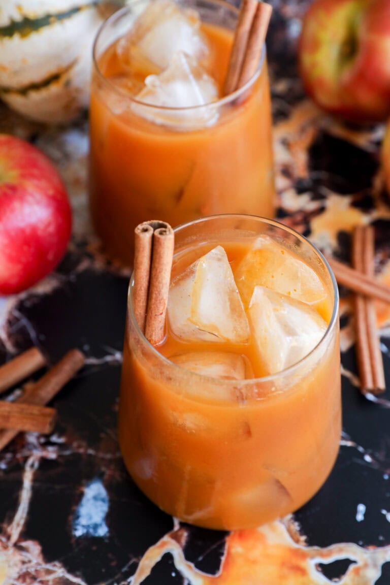 Two glasses of iced apple cider garnished with cinnamon sticks are on a marble surface, surrounded by fresh apples, loose cinnamon sticks for a cozy autumn twist.