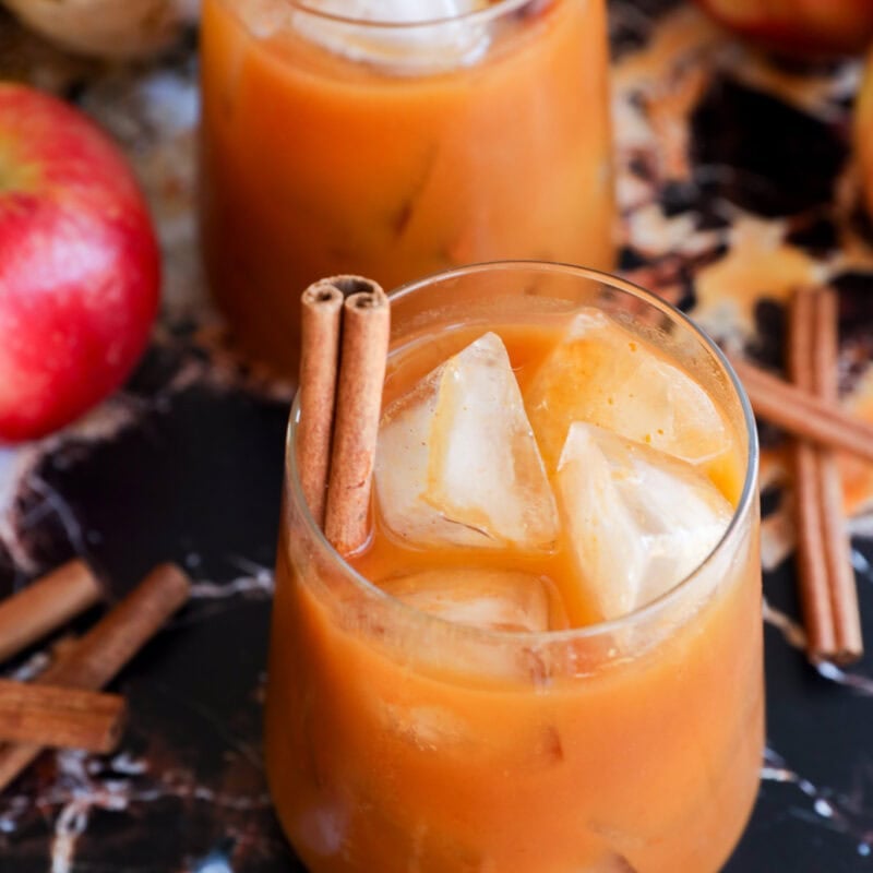 Two glasses of iced apple cider garnished with cinnamon sticks are on a marble surface, surrounded by fresh apples, loose cinnamon sticks for a cozy autumn twist.